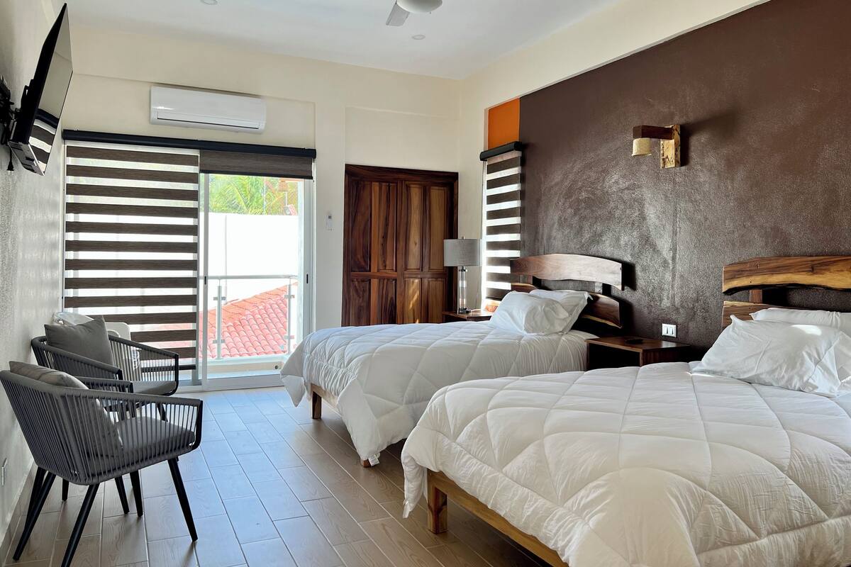A hotel room with two beds, a ceiling fan, an air conditioning unit, a wooden wardrobe, a desk with a lamp, and a view of the outside.