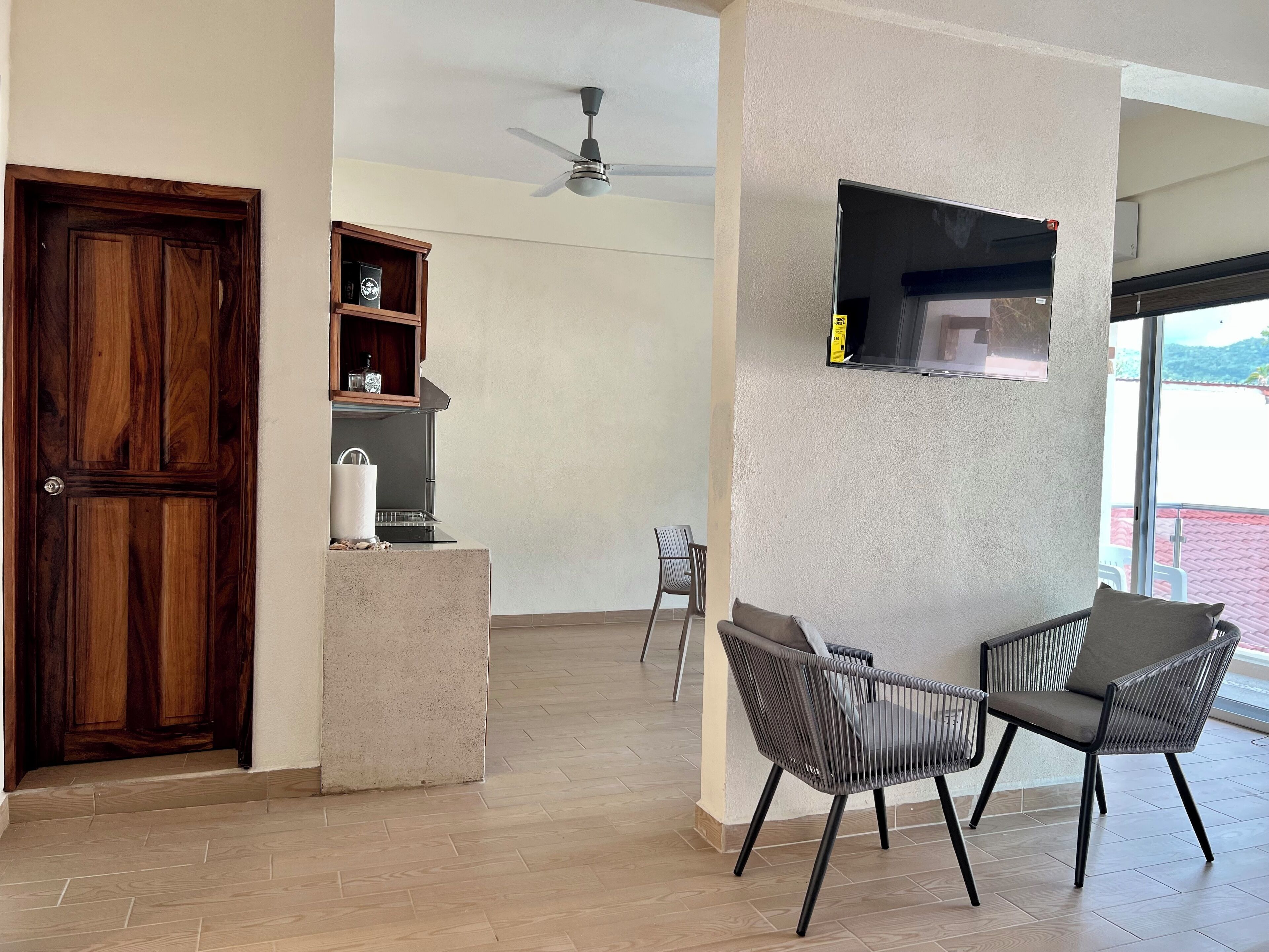 Une pièce spacieuse avec une porte en bois, un ventilateur de plafond, une étagère intégrée contenant des ustensiles de cuisine, un téléviseur à écran plat fixé au mur et deux chaises offrant une vue sur l’extérieur.