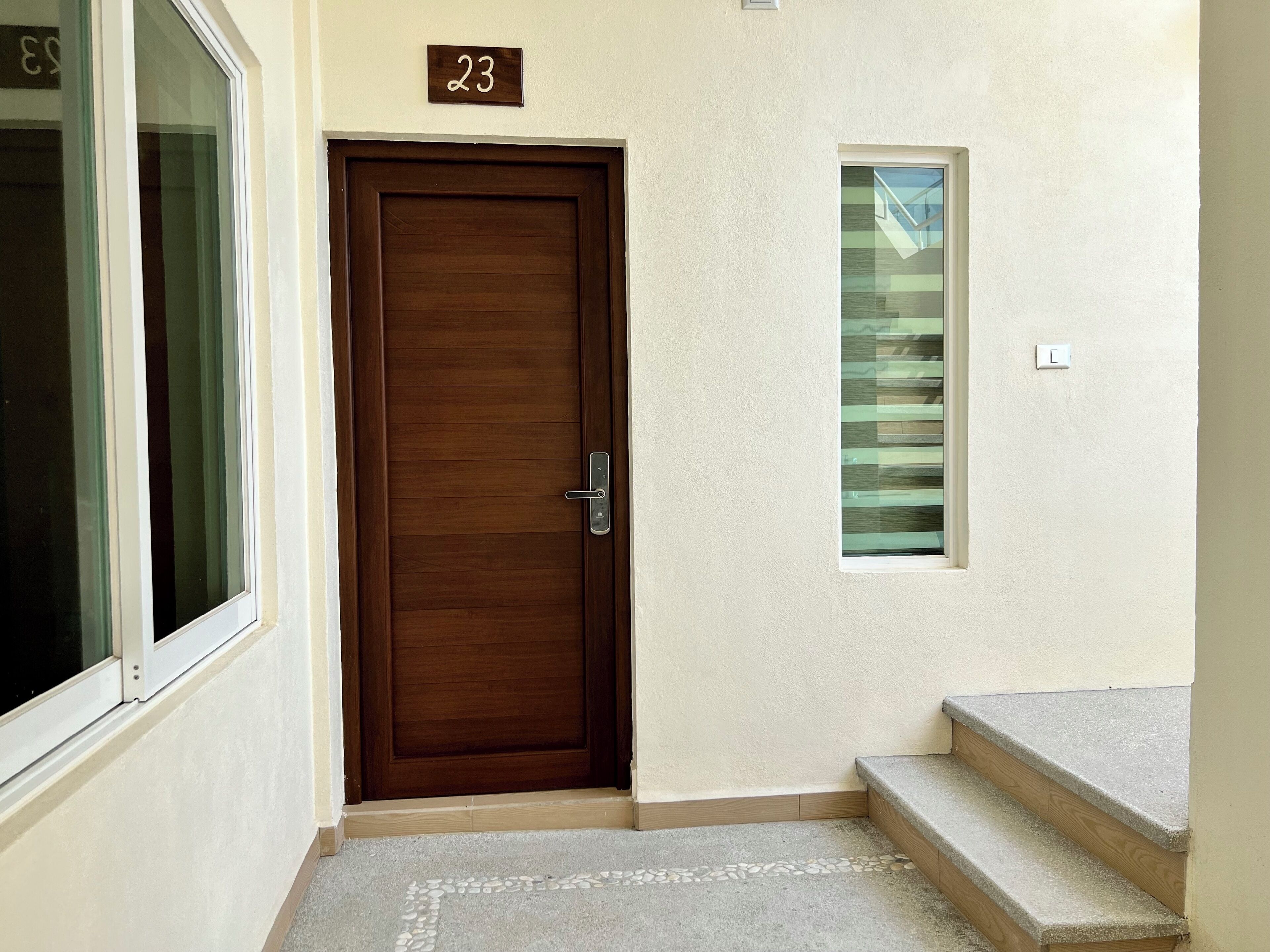 Una puerta de madera con una placa que indica el número 23, un ventanal con persianas y una escalera de hormigón.