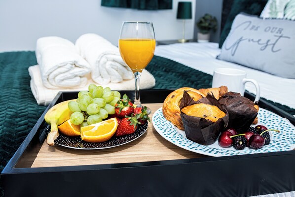 A breakfast tray with fruits, a glass of orange juice, and pastries on a bed with a green blanket and a pillow with "Our Guest" text.