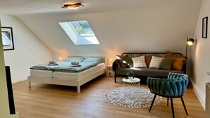 Une chambre à coucher aménagée dans un espace ouvert, avec une lucarne, un lit, un canapé, une chaise et une petite table.