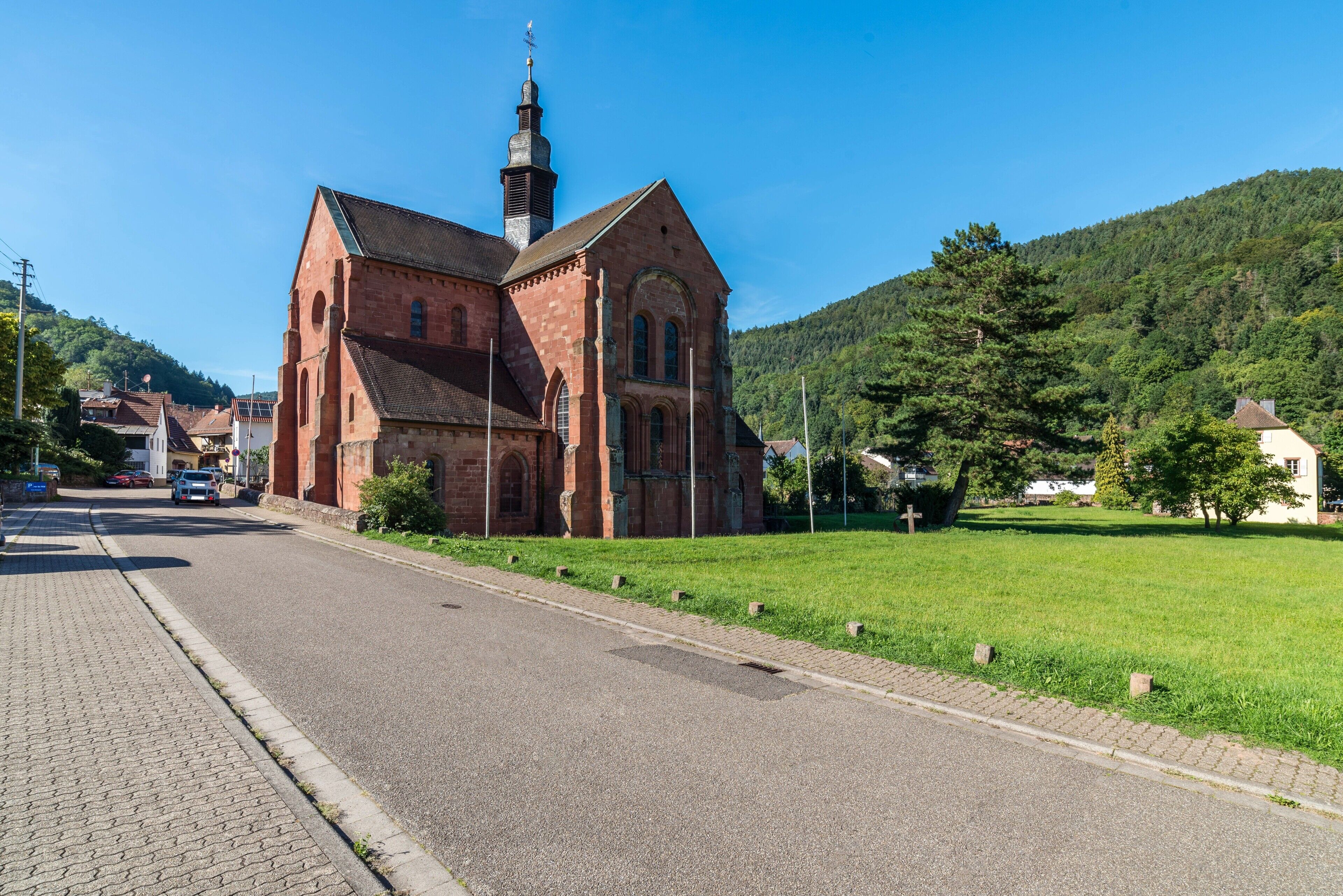 Une église en briques rouges, dotée d’un clocher imposant, une route pavée et un espace verdoyant avec des arbres.
