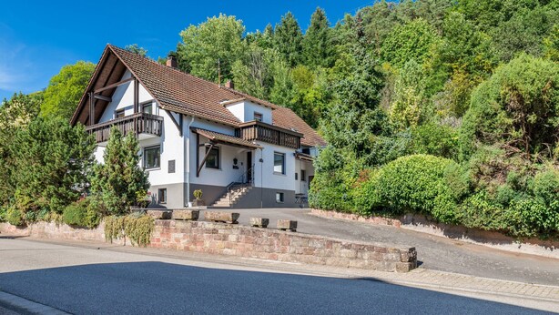 Une maison à deux étages dotée d’un balcon, entourée d’une végétation luxuriante et surplombant un ciel dégagé.