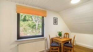 Une pièce avec une fenêtre, une table entourée de chaises, une plante en pot et un radiateur.