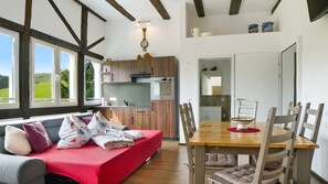 Une pièce chaleureuse avec un canapé rouge, une table à manger et des chaises en bois, une cuisine avec des meubles en bois et de grandes fenêtres donnant sur un paysage verdoyant.