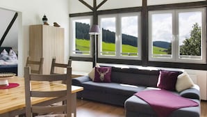 A room with a wooden dining table, a sofa with a view of green fields, and a lamp. - Apartment 'Ferienwohnung 1' with Shared Garden and Wi-Fi (Vöhrenbach)