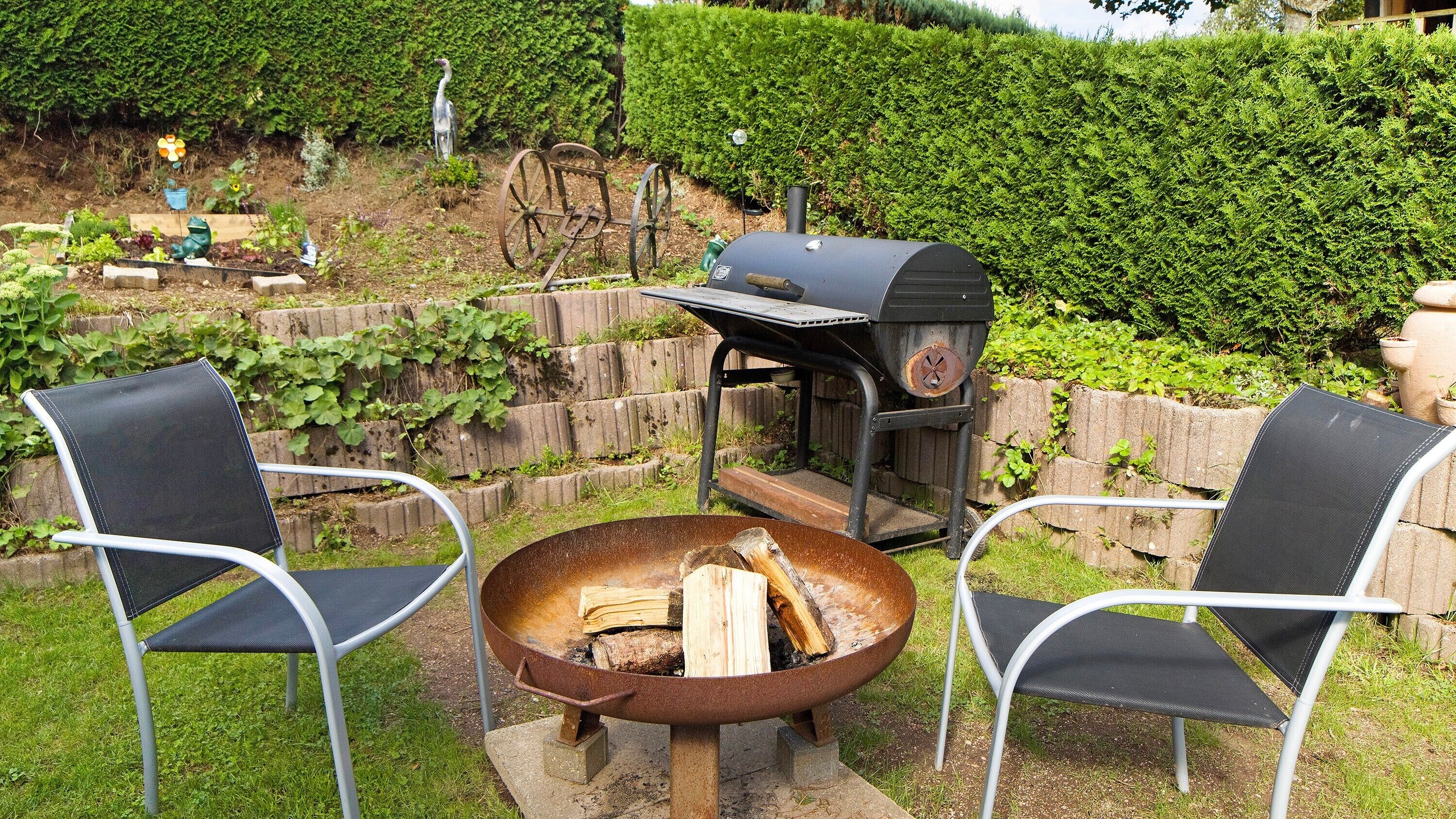 Un espace barbecue extérieur avec un foyer, deux chaises et une haie en arrière-plan.