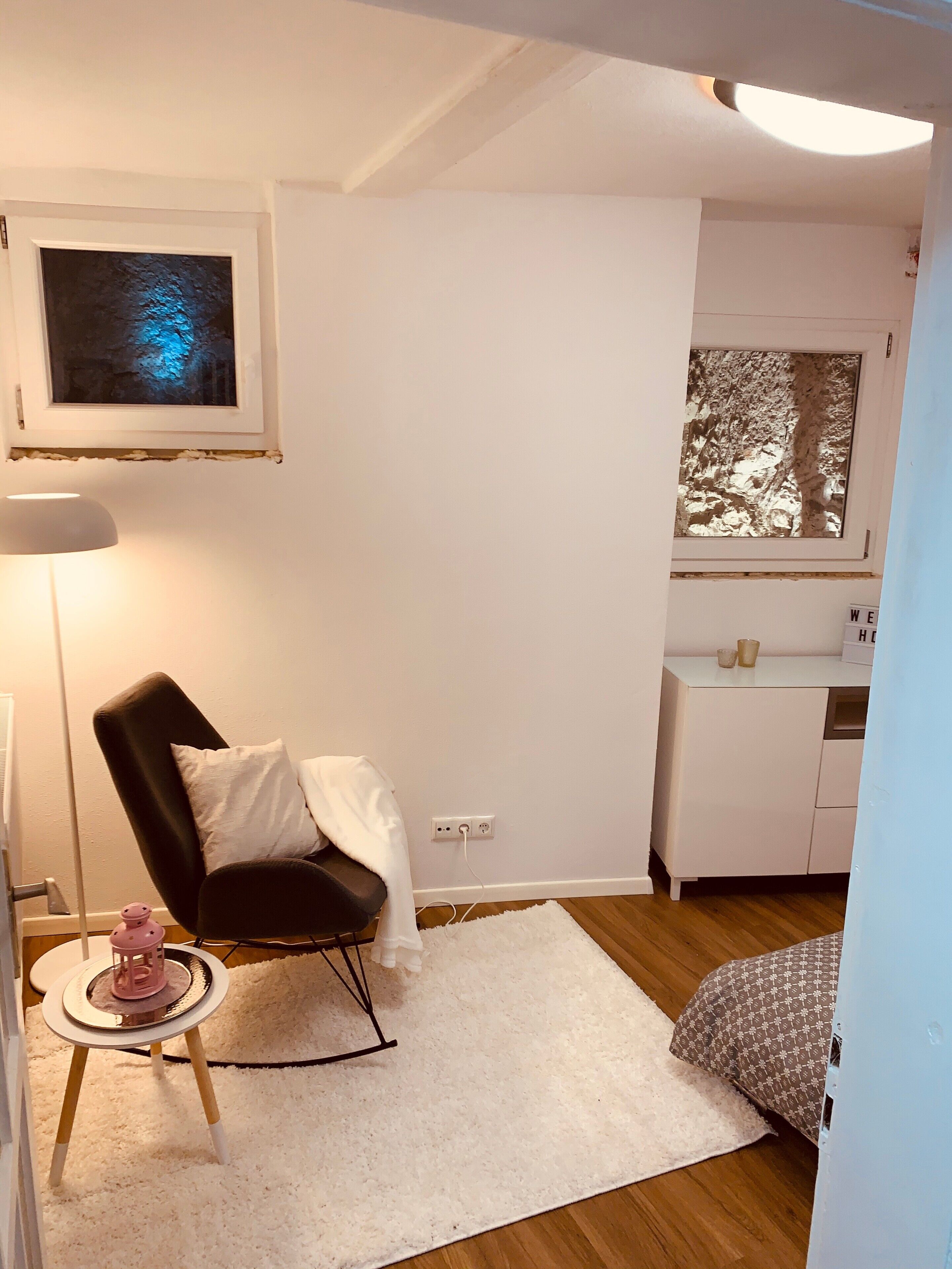 Una habitación pequeña con una silla negra, una alfombra blanca, una mesita con un objeto rosa, una lámpara blanca y una ventana con vistas a árboles.
