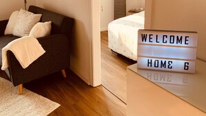 A modern bedroom with a dark sofa, a bedside table, a lamp, and a bed with a 'WELCOME HOME' sign. - Apartment 'Felix Living 7' with Private Terrace and Wi-Fi (Passau)
