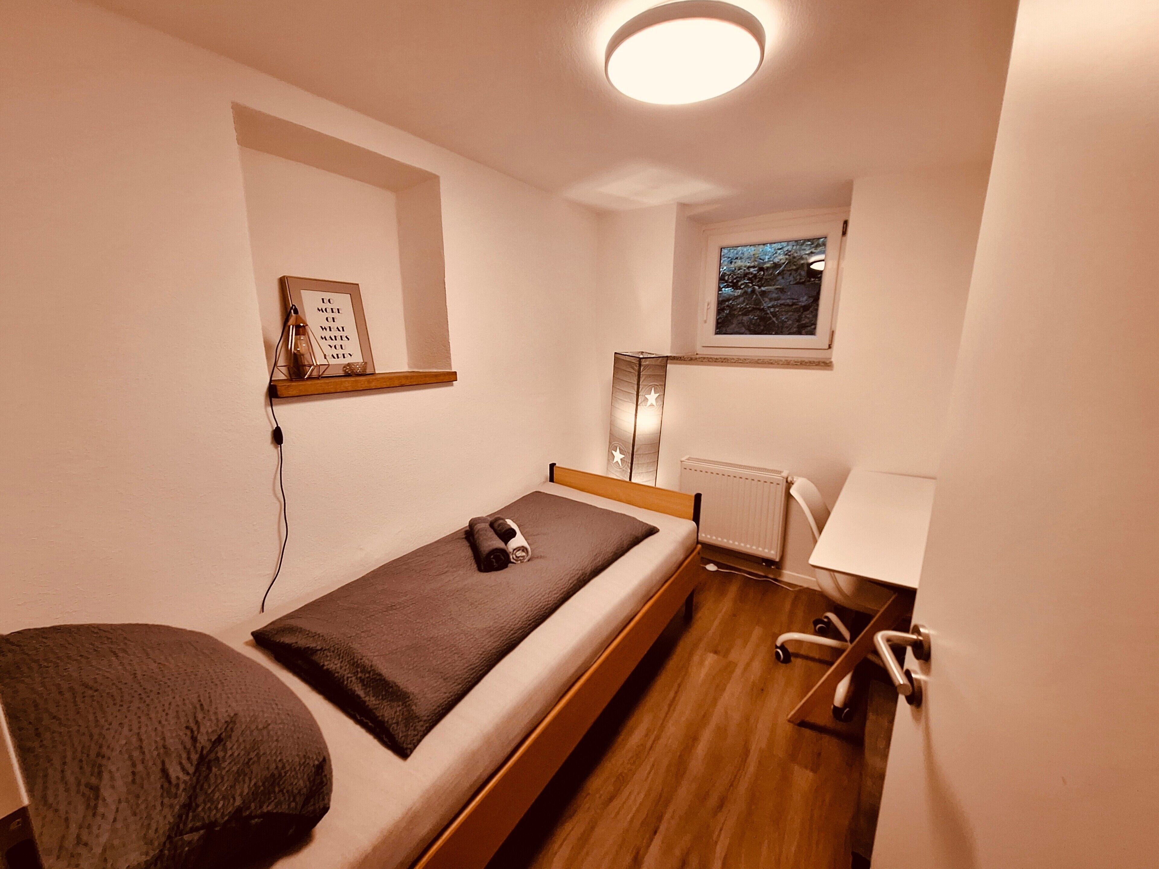 A small, single-bed room with a wooden floor, a bedside table, a lamp, a radiator, and a small window with a view.