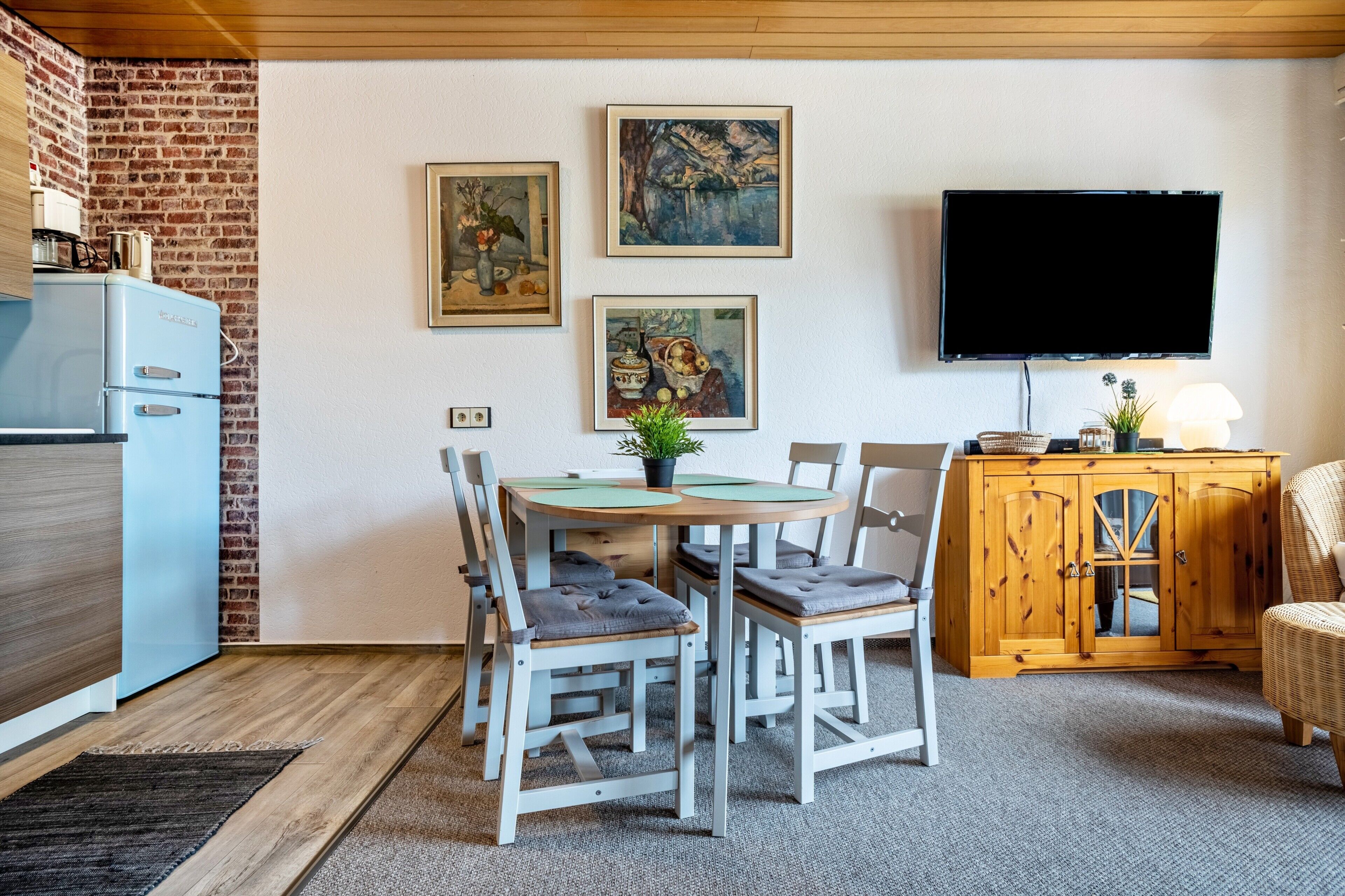 A dining area with a brick wall, a blue refrigerator, a wooden cabinet, and a TV mounted on the wall.