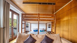 Ein Zimmer mit Holzboden, einem Bett mit blau-weißer Bettwäsche, einem beigen Sofa mit zwei Kissen und einem großen Fenster mit Vorhängen.
