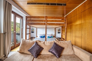 Ein Zimmer mit Holzboden, einem Bett mit blau-weißer Bettwäsche, einem beigen Sofa mit zwei Kissen und einem großen Fenster mit Vorhängen.