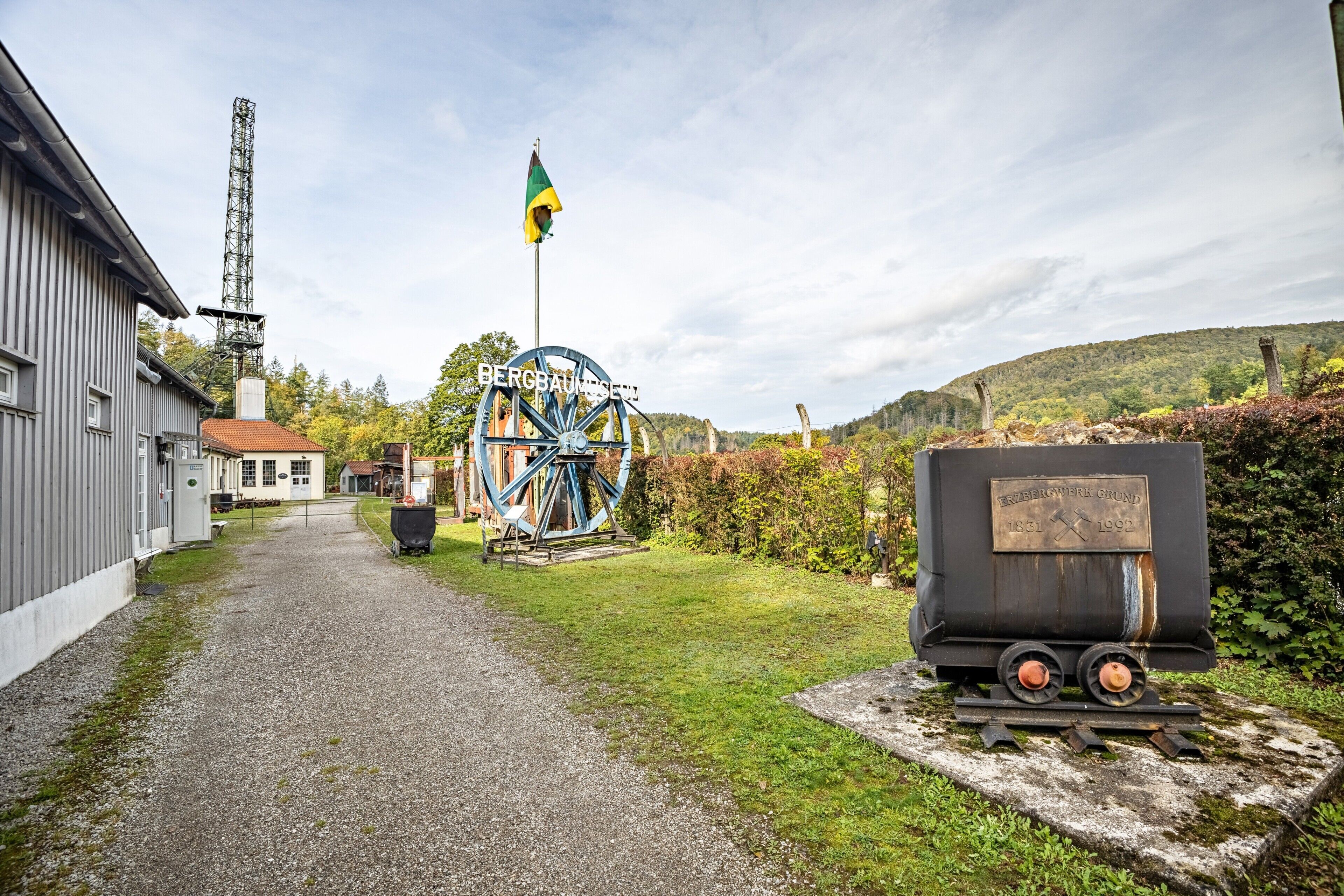 A gravel path leading to a building with a large water wheel and a flagpole.
