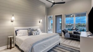 Un dormitorio con una cama grande, dos luces fijadas a la pared, un ventilador de techo, vistas al exterior a través de amplias ventanas y un taburete pequeño.