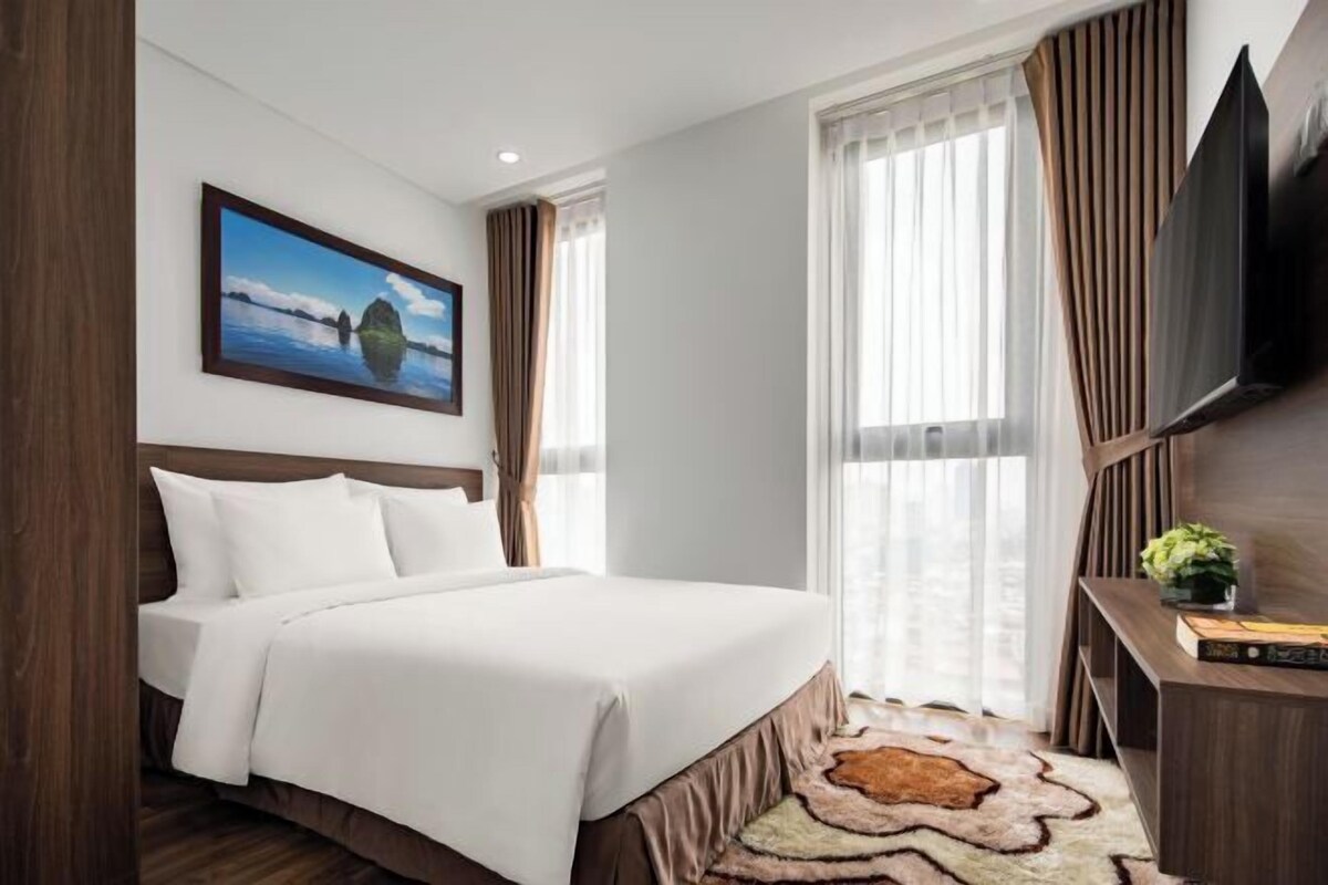 Une chambre d’hôtel avec un lit, un téléviseur fixé au mur, un tableau et une fenêtre avec des rideaux.