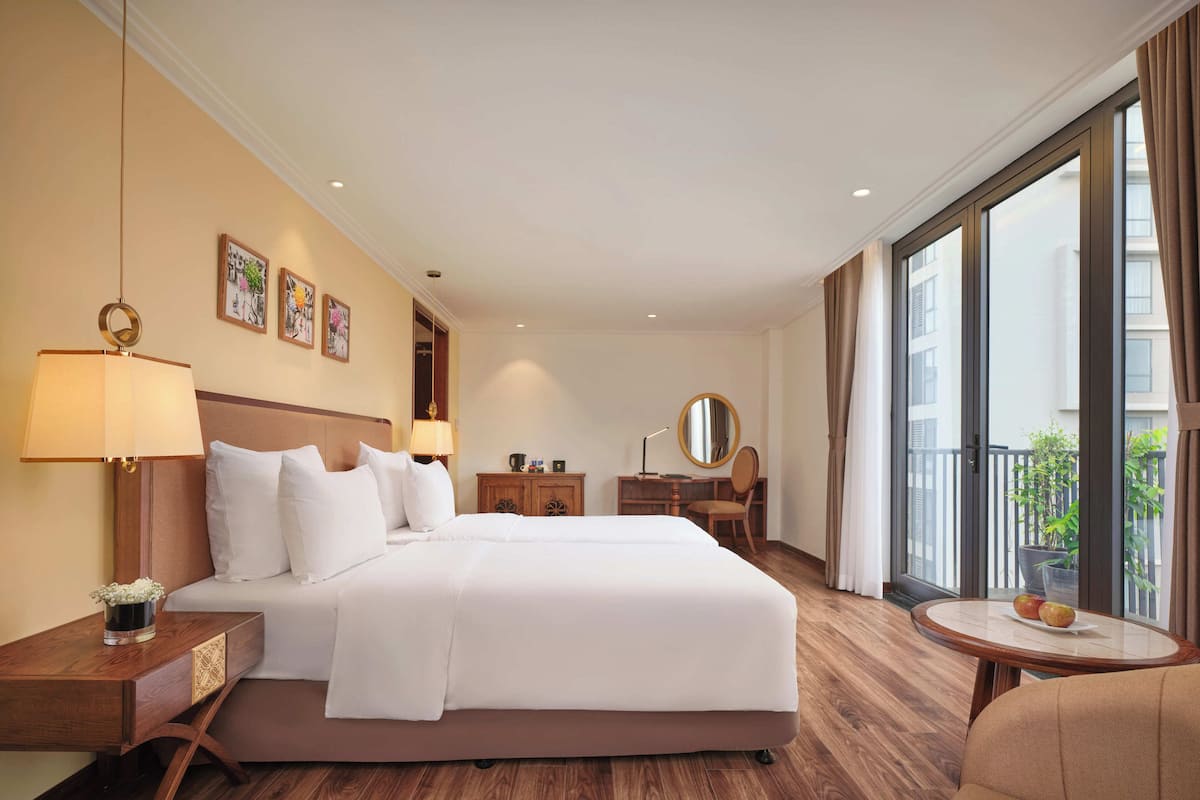 Een hotelkamer met een groot bed, een bureau, een stoel, een ronde tafel met fruit en een balkon met een plant.