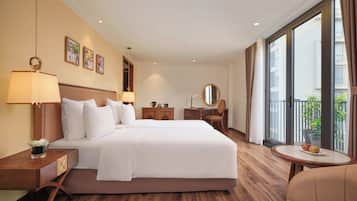 A hotel room with a large bed, a desk, a chair, a round table with fruit, and a balcony with a plant.