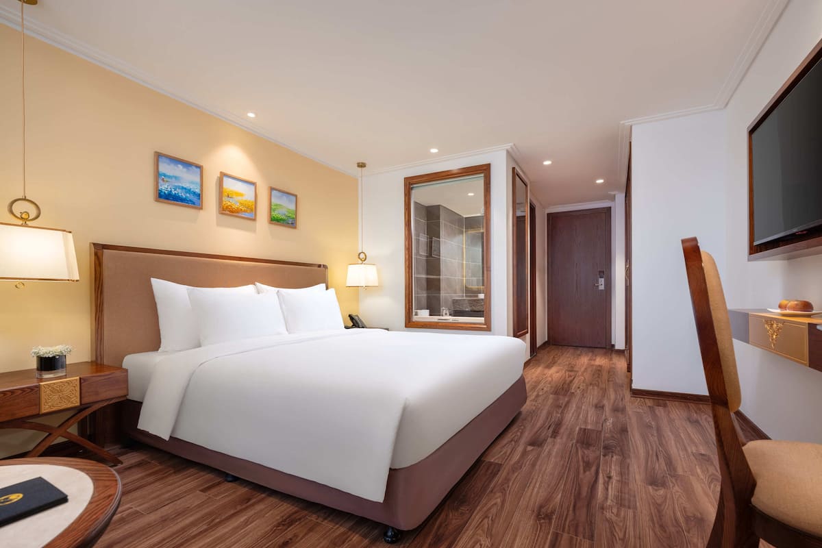 Hotelkamer met een groot bed, houten vloer, een bureau met stoel, een aan de muur bevestigde televisie en een badkamer door een glazen deur zichtbaar.