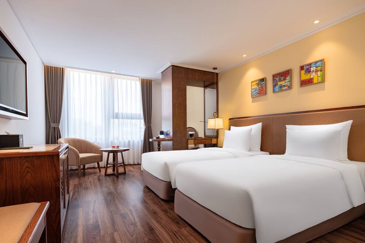Een hotelkamer met houten vloer, een bureau met computer, twee bedden, een stoel, een klein tafeltje, een lamp en een grote spiegel.
