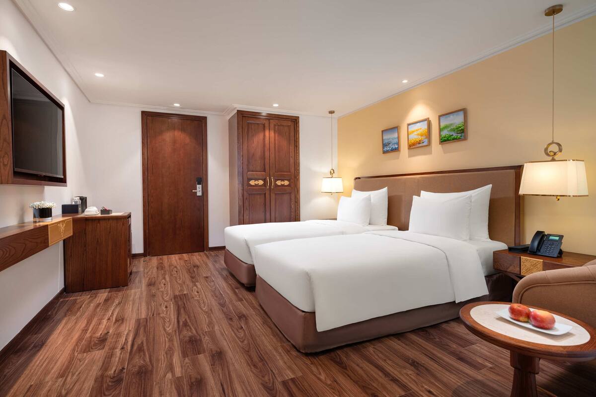 Een hotelkamer met een tweepersoonsbed, een houten garderobe, nachtkastje, lamp en een klein tafeltje met fruit.