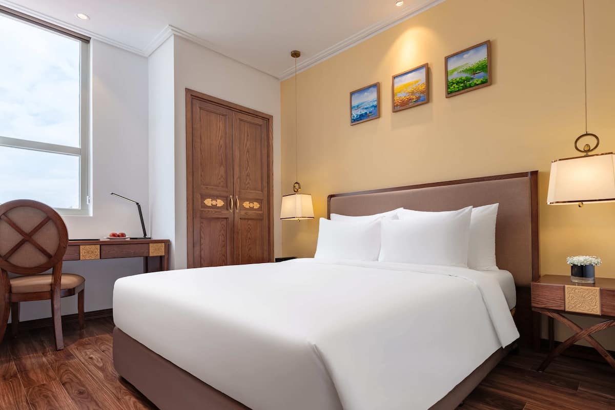 Een hotelkamer met een groot bed, een bureau met stoel, een houten kledingkast en drie schilderijen aan de muur.
