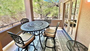 A covered patio with a round table and four chairs, a view of trees and a building in the background. - Canyon Vista 🌵3br Retreat: Views, Hiking, Birds, Heated Pools, Spas, Court, Gym (Tucson)