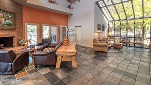 A spacious living area with a fireplace, leather seating, and a large window offering a view of the outdoors. - Canyon Vista 🌵3br Retreat: Views, Hiking, Birds, Heated Pools, Spas, Court, Gym (Tucson)