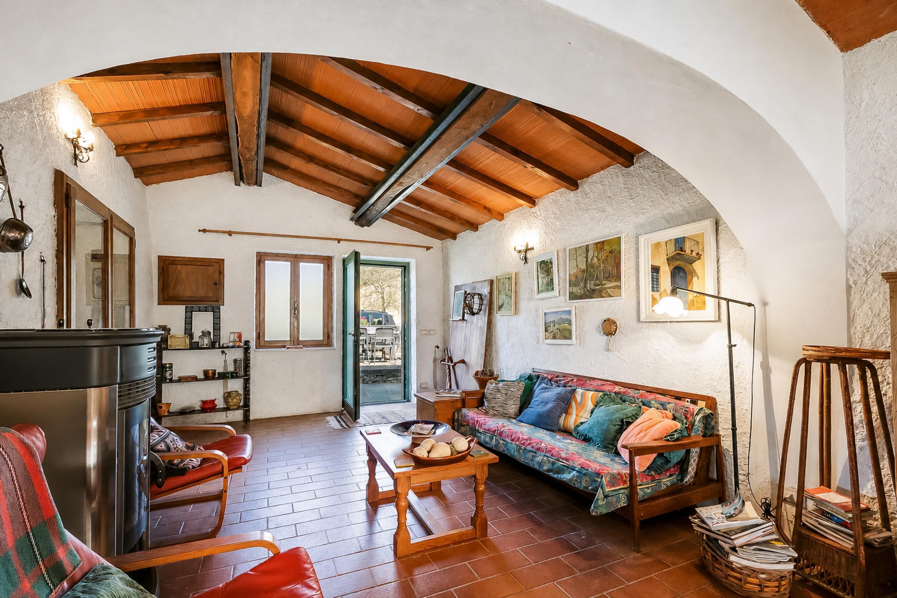Ein rustikales Interieur mit Holzbalkendecke, einer Couch mit bunten Kissen, einem Esstisch und einer Küchenzeile mit Kühlschrank und Regalen.