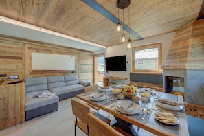 A modern living room with a wooden ceiling, a grey sofa, a dining table set with plates and glasses, and a fireplace. - Wood House Livigno Ski in - Ski out 10m, Livigno, Italy (Livigno)