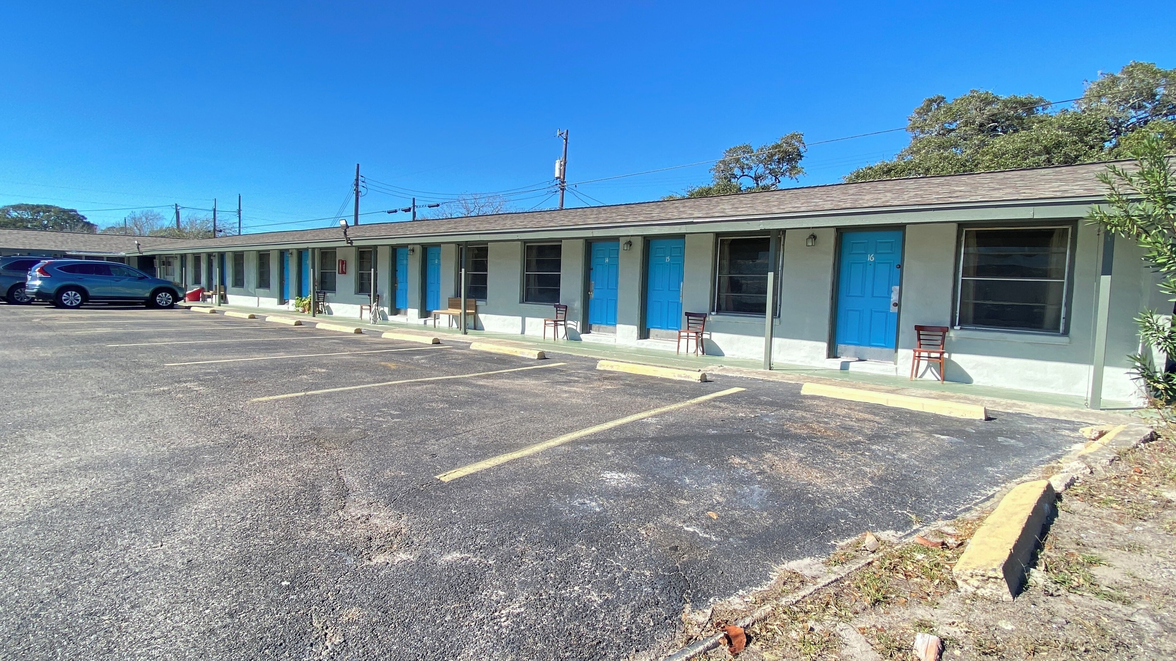 Un motel avec une rangée de chambres, chacune dotée d’une porte bleue et d’une fenêtre. Un parking se trouve devant le motel.