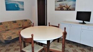 A dining area with a round table, wooden chairs, a patterned sofa, a television on a cabinet, and two wall-mounted pictures of ocean scenes.