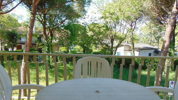 A round table with a single chair in the foreground, a white railing, and a view of a green landscape with trees and buildings in the background.