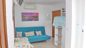 A room with a blue sofa, floral cushions, a painting of a sunset, a wooden door, and a shelf with a patterned cloth. - Apartment Malva in Conil (Conil de la Frontera)