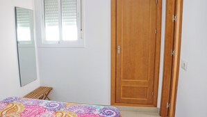 A bedroom with a wooden door, a bed with a colorful bedspread, a small wooden stool, and a window with blinds. - Apartment Malva in Conil (Conil de la Frontera)