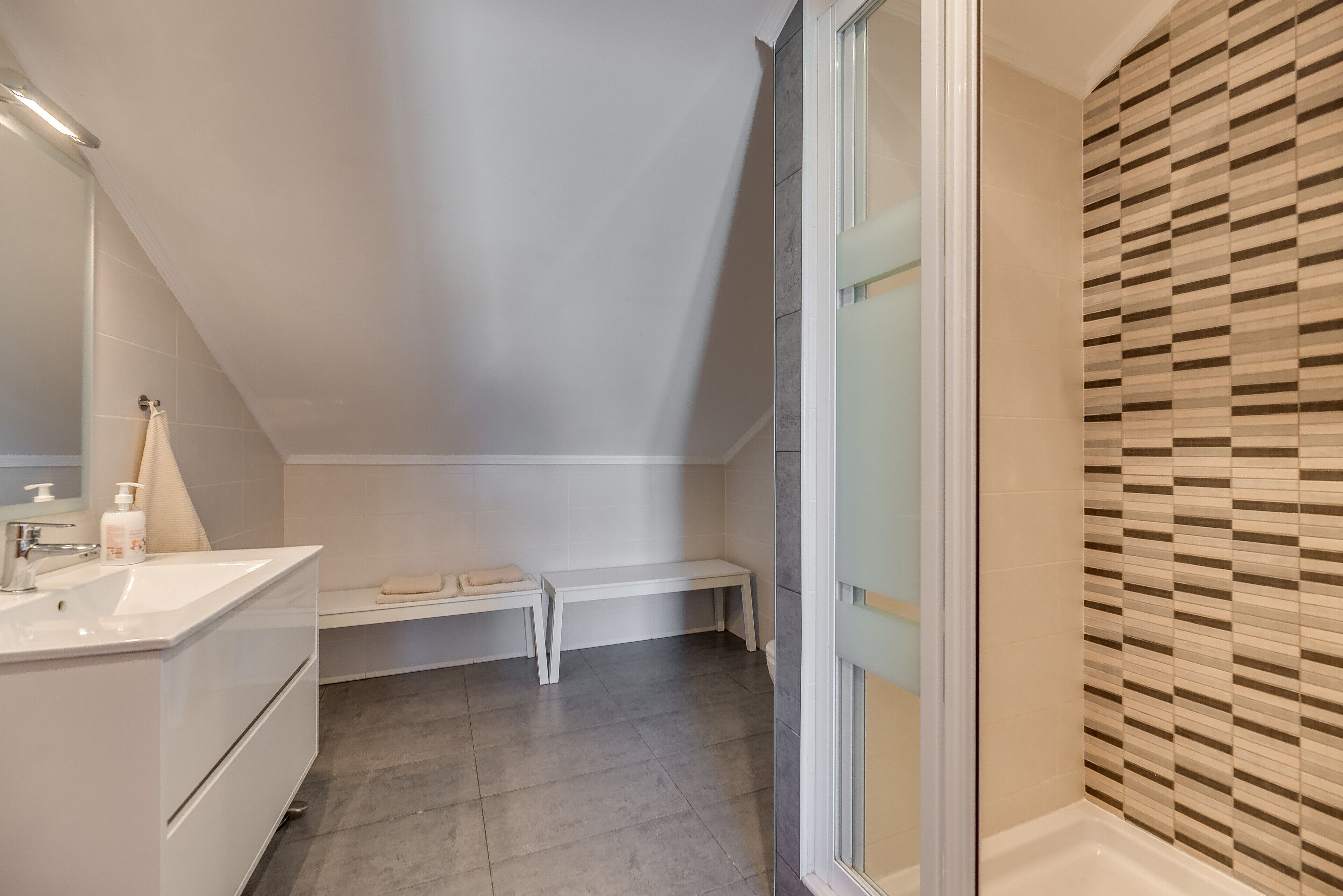 A modern bathroom with a white vanity, a bench, and a shower area.