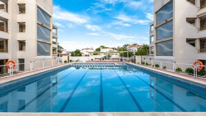 Uma piscina com água azul cristalina, rodeada por edifícios modernos com varandas, e um céu claro acima.