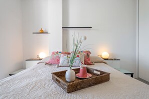 A neatly made bed with a white quilt, a wicker tray with a vase and a checkered cup, and decorative pillows with a light bulb on each nightstand.
