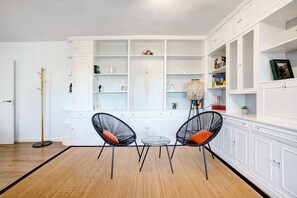 Una habitación con un armario blanco, dos sillas negras con cojines naranja, una mesita redonda y una lámpara de pie.