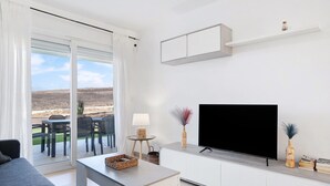 Ein modernes Wohnzimmer mit Fernseher, weißen Schränken, einem Essbereich mit Stühlen und Blick durch die Glastür.
