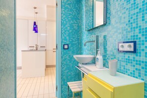 Un baño con pared de mosaico azul, lavamanos blanco, mueble amarillo y lámpara colgante azul.