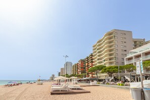 Une plage avec des transats, des parasols et une promenade bordée de bâtiments en arrière-plan.