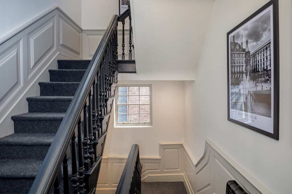 Un escalier avec des marches recouvertes d’une moquette sombre et une rampe noire, menant à un palier doté d’une fenêtre.