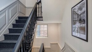 Un escalier avec des marches recouvertes d’une moquette sombre et une rampe noire, menant à un palier doté d’une fenêtre.