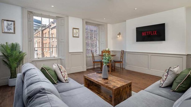 Un salon avec un canapé gris, une table basse en bois et un téléviseur fixé au mur qui affiche le logo « NETFLIX ».