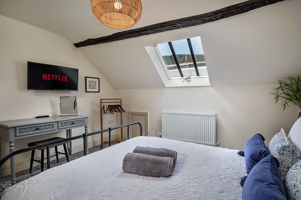 Une chambre à coucher avec un lit, un bureau avec un ordinateur, un téléviseur affichant le logo « NETFLIX », un puits de lumière et un radiateur.