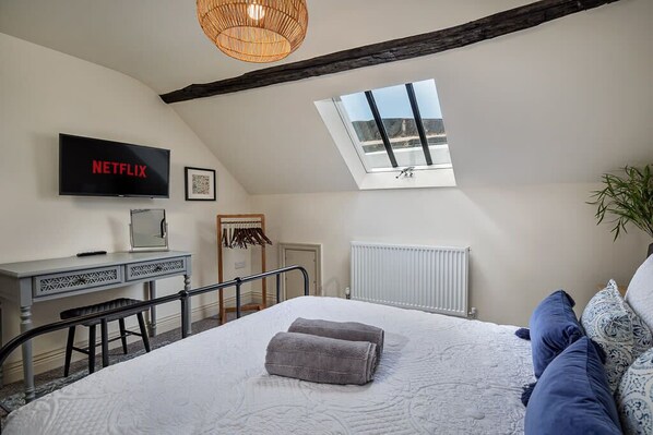 A bedroom with a bed, a desk with a computer, a television displaying 'NETFLIX,' a skylight, and a radiator.