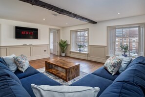 A living room with a large TV mounted on the wall, a wooden coffee table, blue sofas, and a view through the windows.