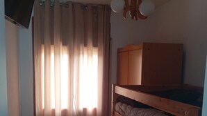 A room with a bunk bed, a wooden wardrobe, a television mounted on the wall, and a window with curtains.
