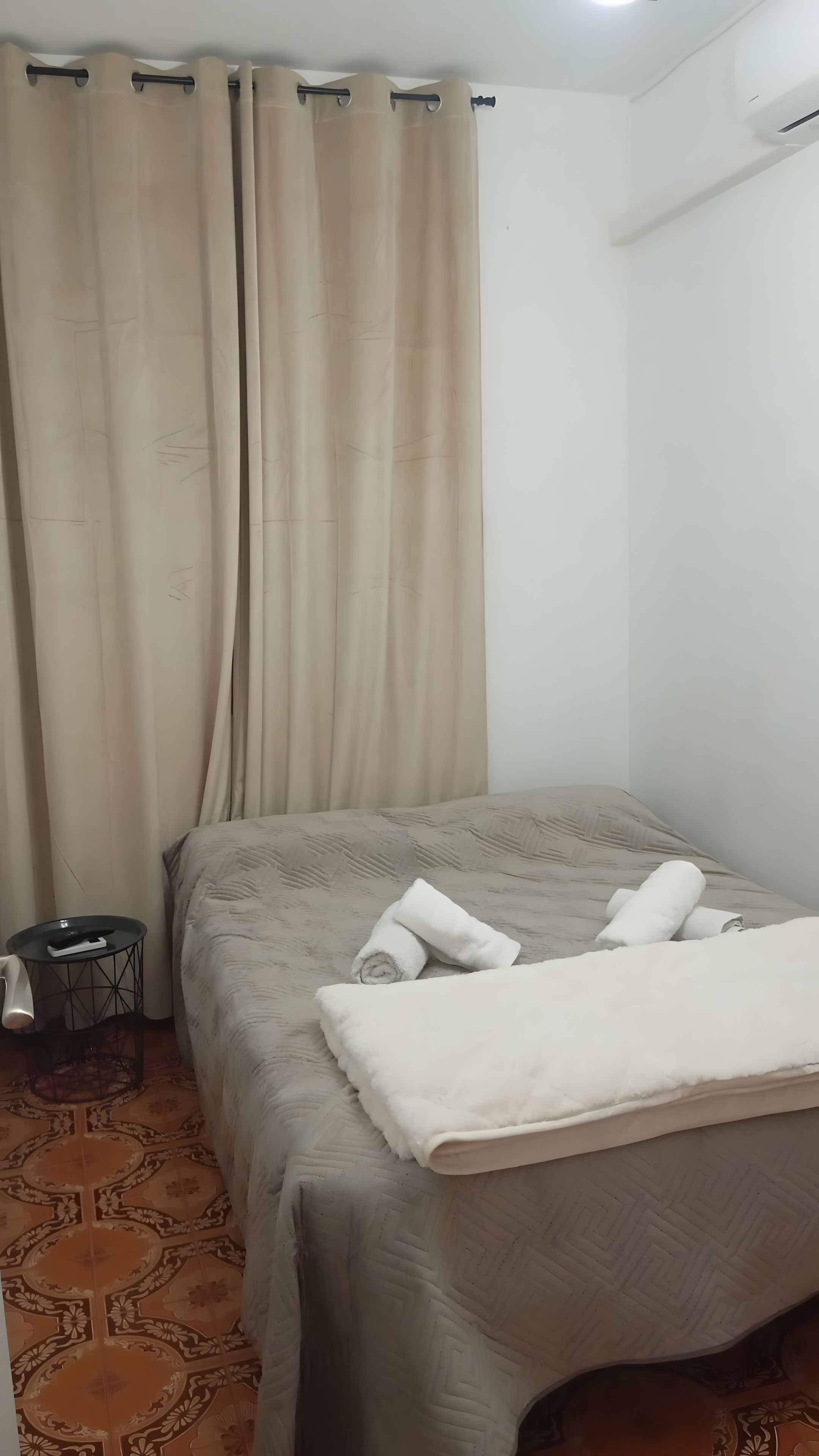 A bedroom with a bed, beige curtains, a small side table, and a patterned floor.